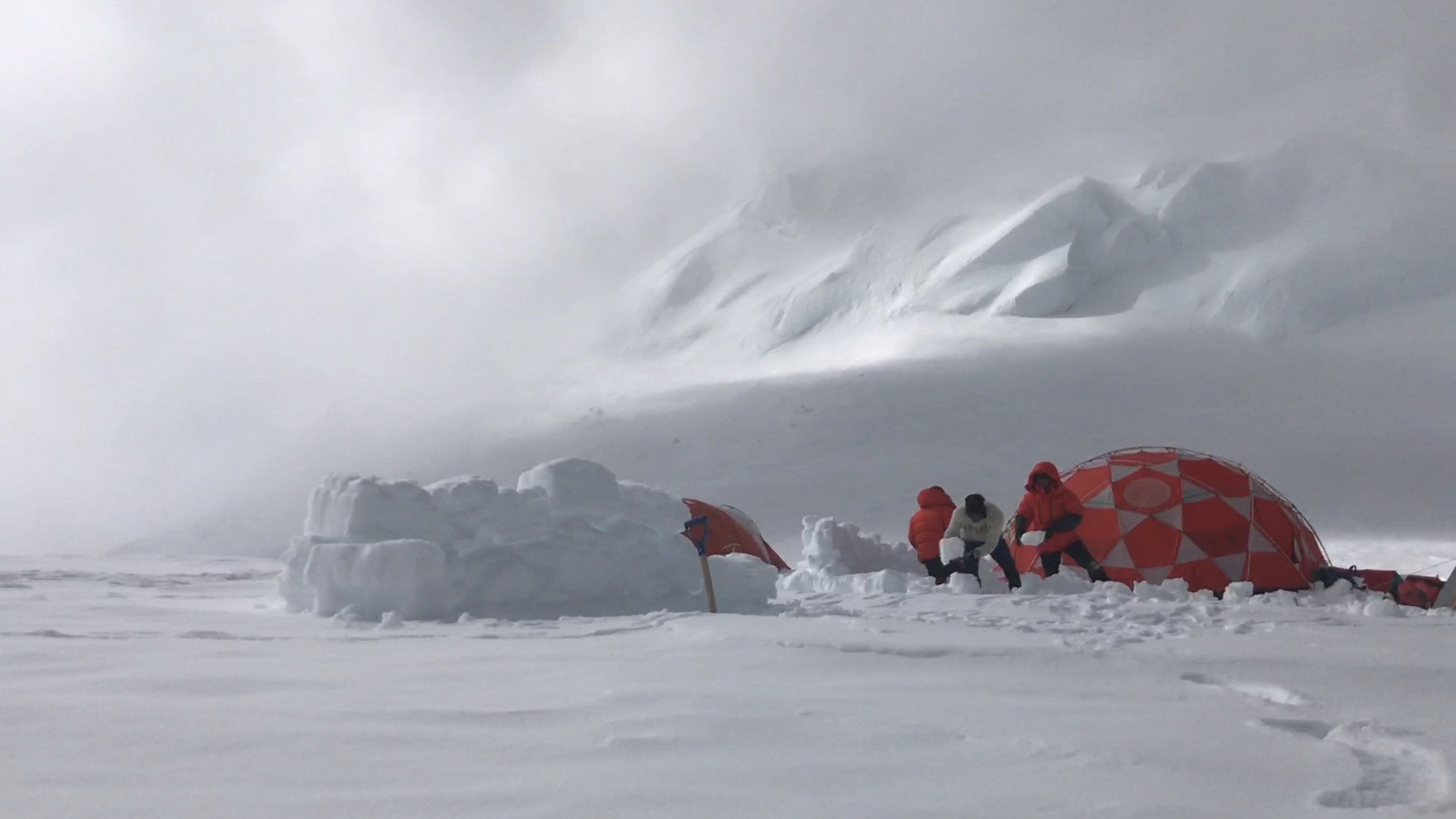 Expedition members construct a snow brick wall to shield camp structures from heavy winds on July 17, 2019. Photo credit: Forrest Schoessow.