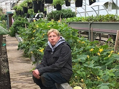 Baldy-Hughes-Vernon-Brendan - blonde-haired man in grey hoodie and jacket sits outside on edge of raised bed of vegetables Baldy-Hughes-Vernon-Brendan