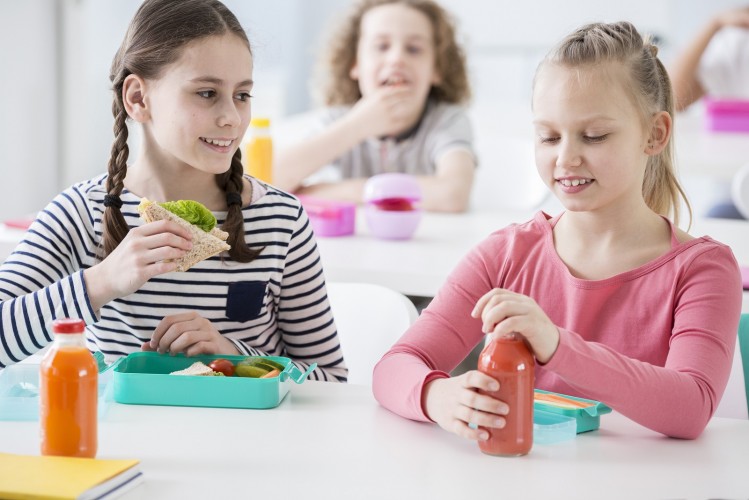Front View Of Two Junior Girls In A School Cafeteria During Lunch Break. One Holding A Wholewheat Sa