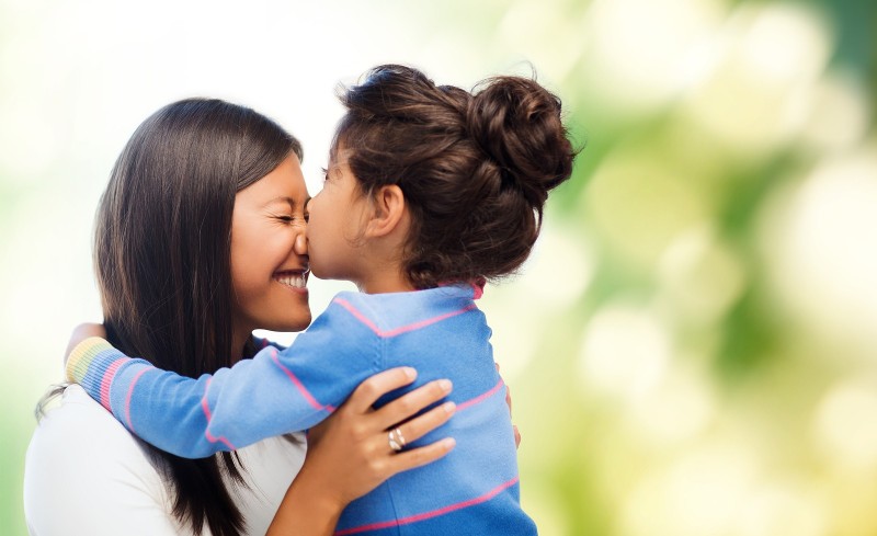 family, children and happy people concept - happy little girl hugging and kissing her mother over gr