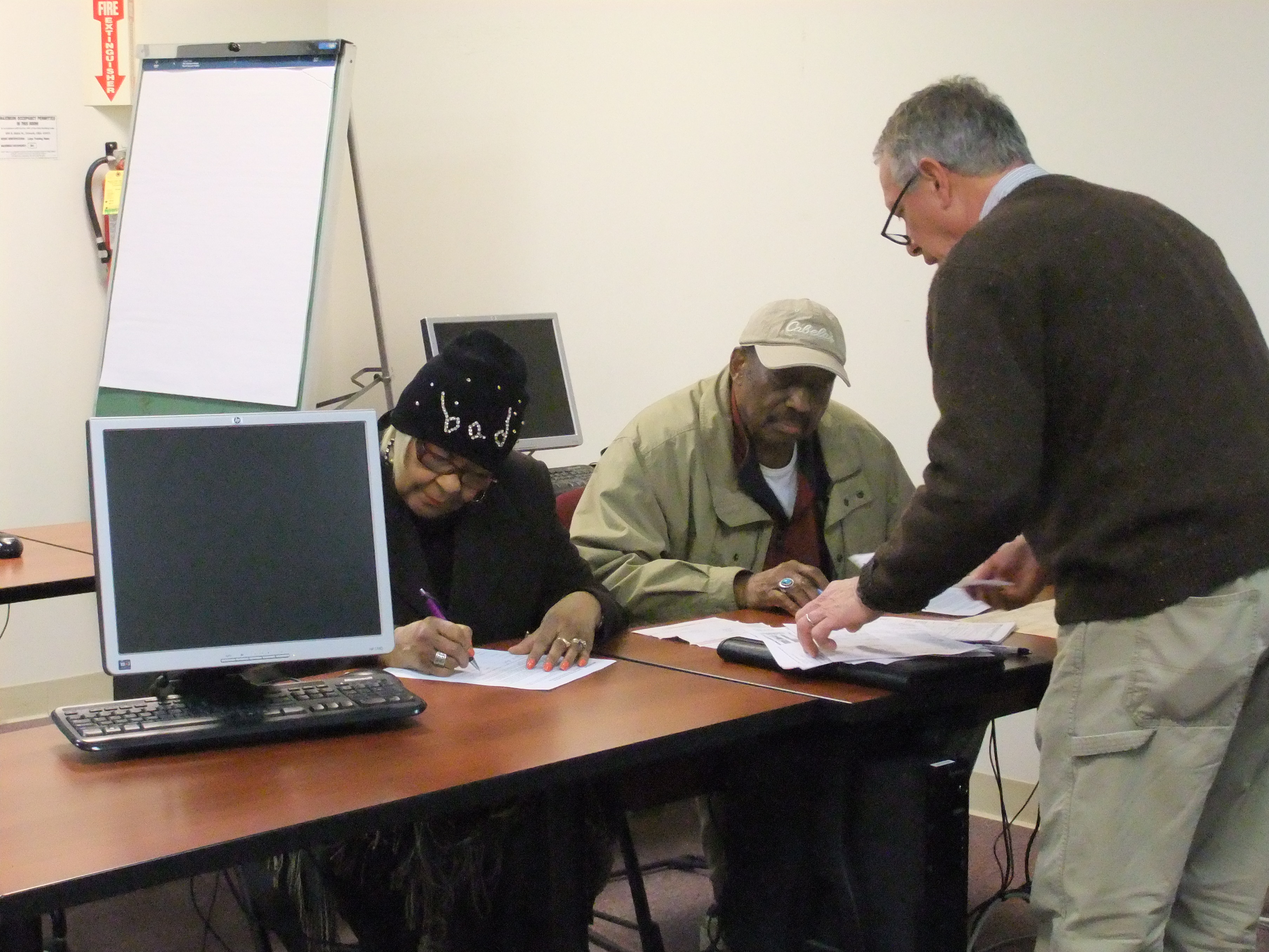 two people sitting at a table with paperwork in front of them they are with pens in their hands filling out paperwork as another man is standing in front of them helping them with their paperwork.