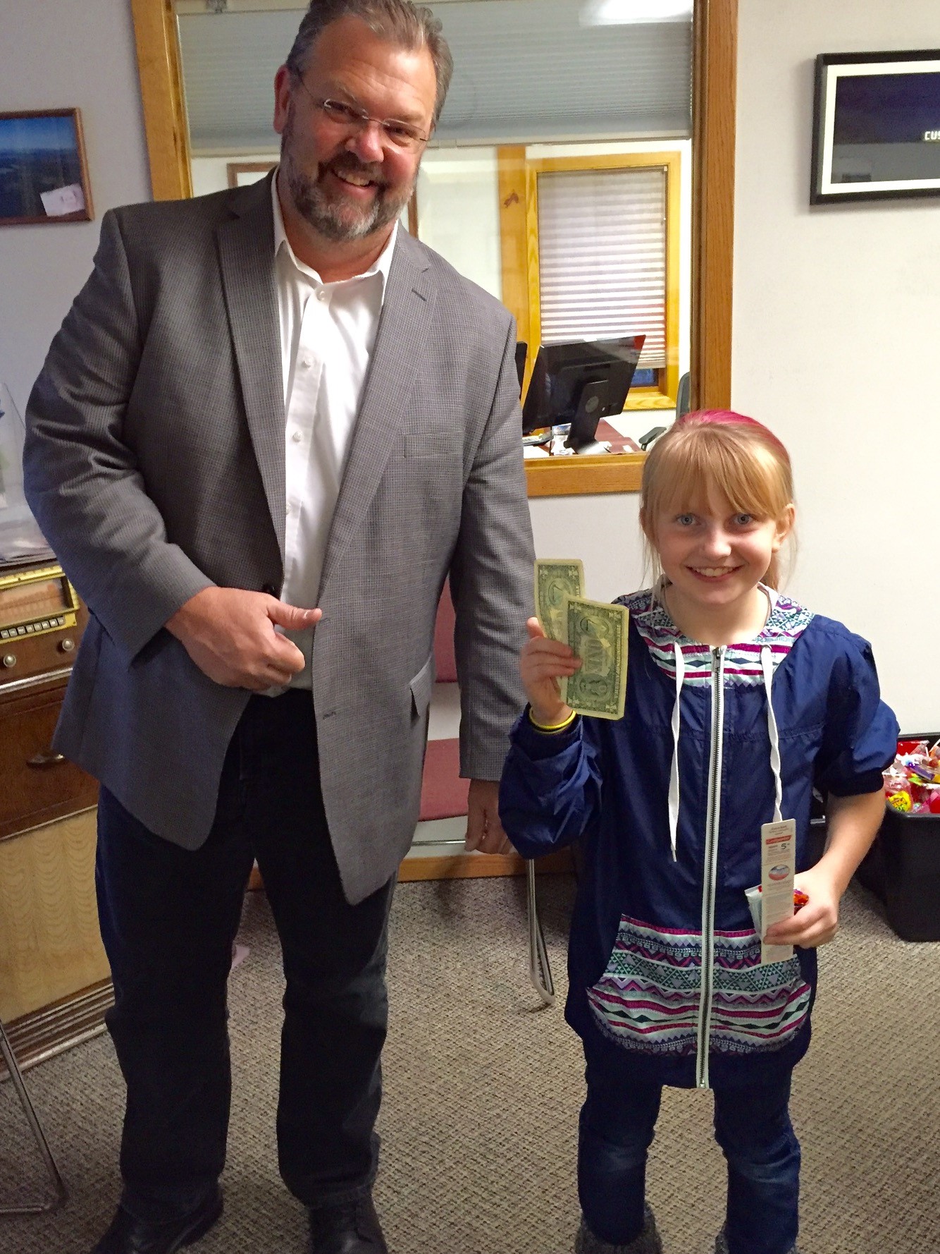 Smiling man standing next to a smiling young girl who is holding up two one dollar bills in her hand