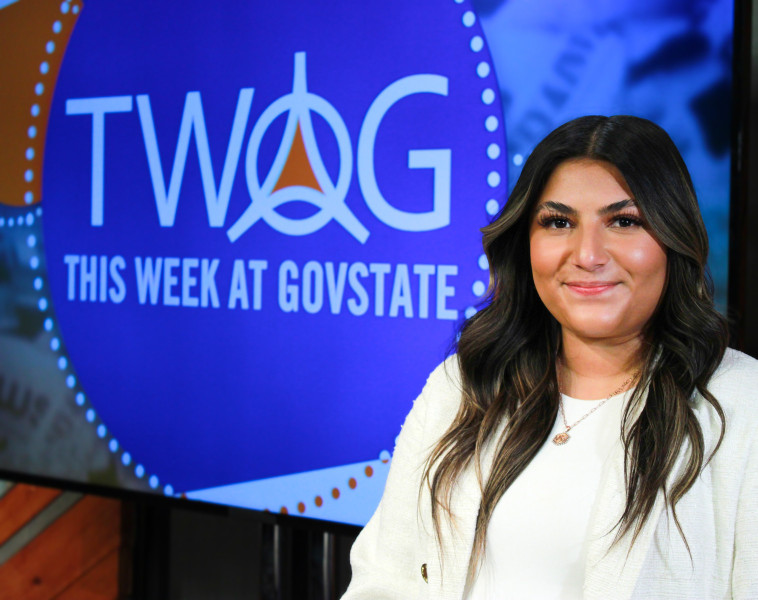 A smiling woman standing in front of the This Week at GovState news show studio screen