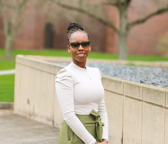 A smiling woman in a cream long sleeve shirt and olive pants standing outside on a patio