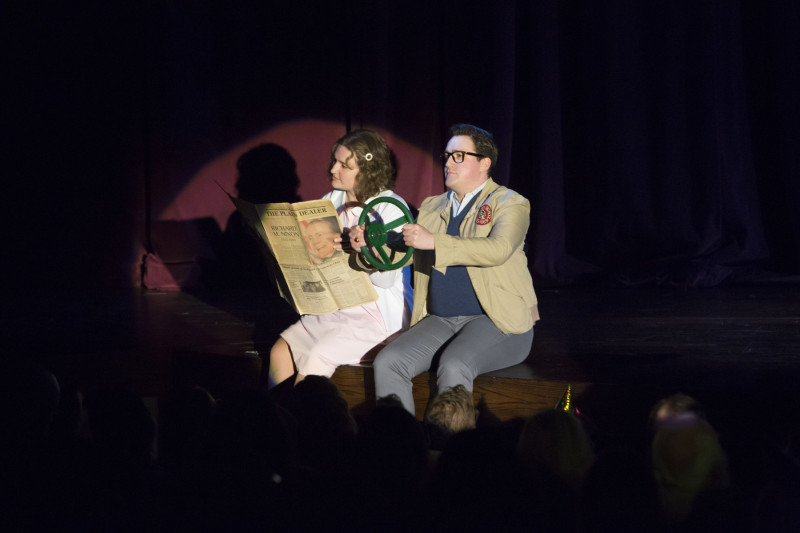Two people in costume on a stage acting out driving a car