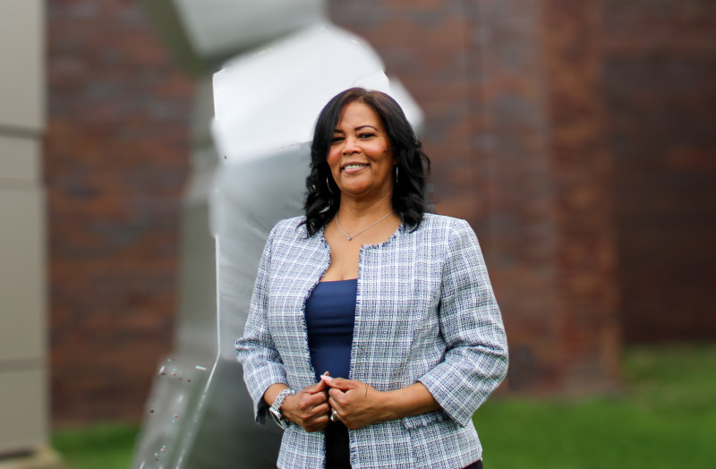 A smiling woman in blue shirt and gray plaid blazer standing outside in front of a geometric sculpture