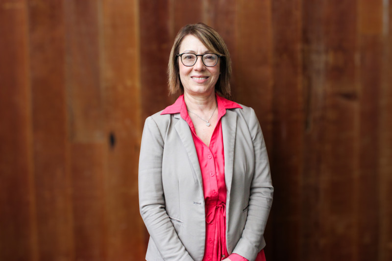 A smiling woman in a pink shirt and gray blazer standing in front of a wood paneled wall
