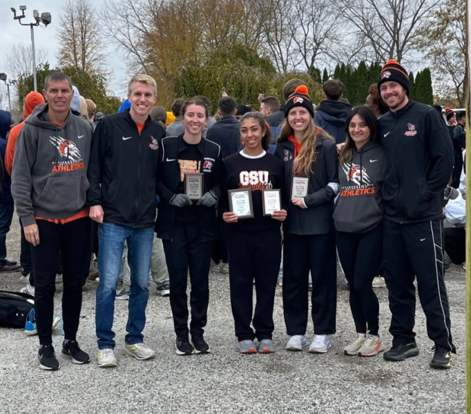 Three Governors State University women's cross country runners smiling while holding small championship plaques and standing with the four member coaching team