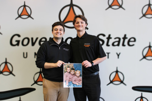 Two brothers smiling for a photo while holding a childhood photo of themselves