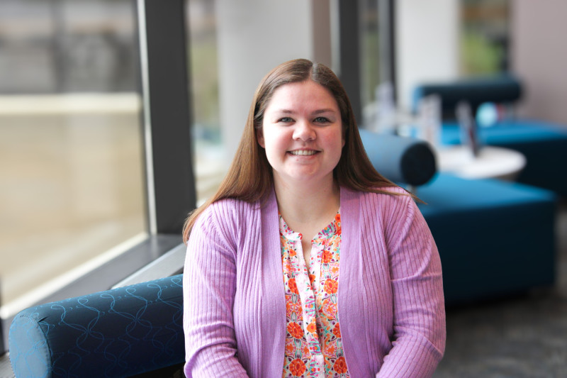 A smiling woman in a lilac sweater sitting on a blue  couch in front of a wall of windows