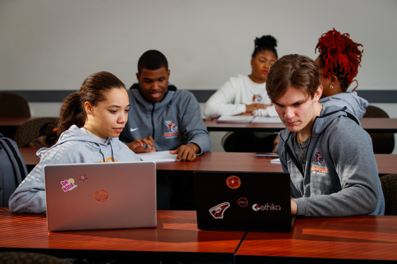 Five college students sitting at tables working on laptops