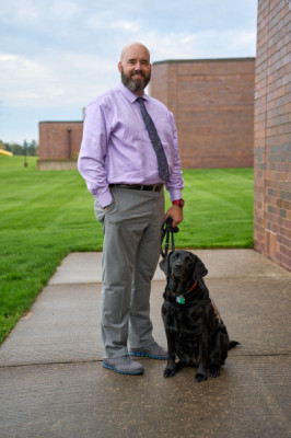 Portrait shot of a smiling man in lavender shirt and gray pants with his black service dog standing outside Governors State University