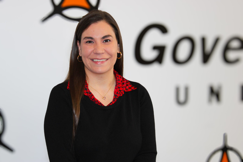 A smiling woman in a black sweater standing in front of Governors State University's mural