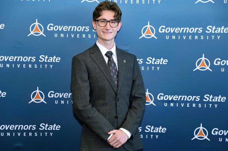 Portrait shot of a smiling man wearing a dark gray suit with white shirt, navy tie and glasses
