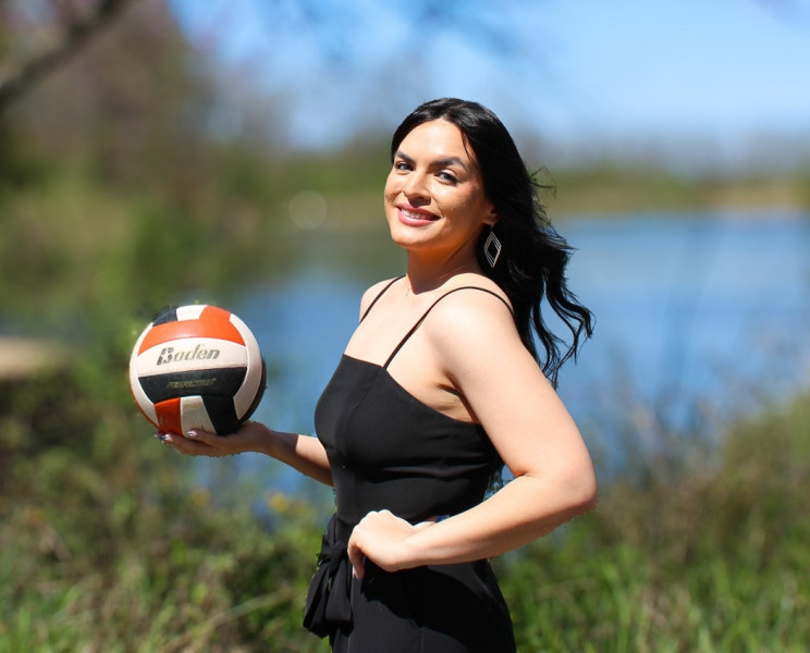 A smiling woman wearing a black dress holding a volleyball in front of Governors State University lake