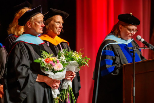 Two women in doctoral academic regalia receiving flowers from another woman in academic regalia