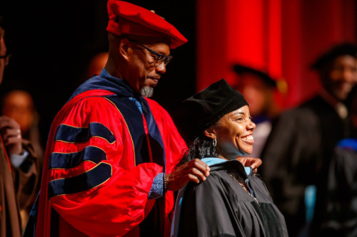 A smiling woman in doctoral regalia receiving her hood from a man in academic regalia