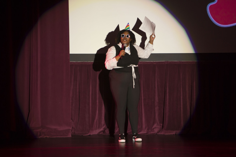A woman wearing a black pantsuit speaking into a microphone on a theater stage