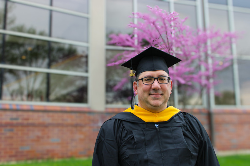 A man in black graduation cap and gown standing outside in front of a pink flowered tree