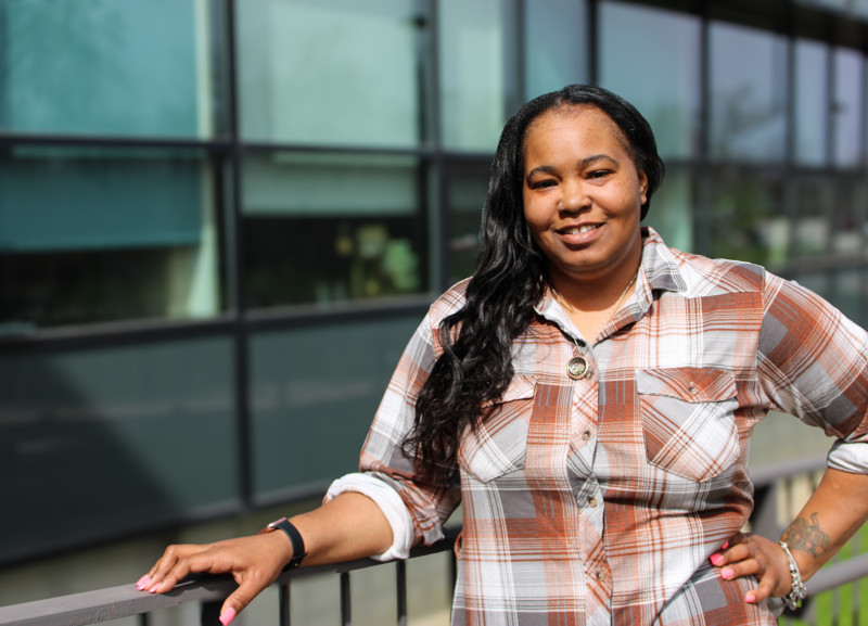 A smiling woman wearing a plaid shirt leaning against a metal guard rail