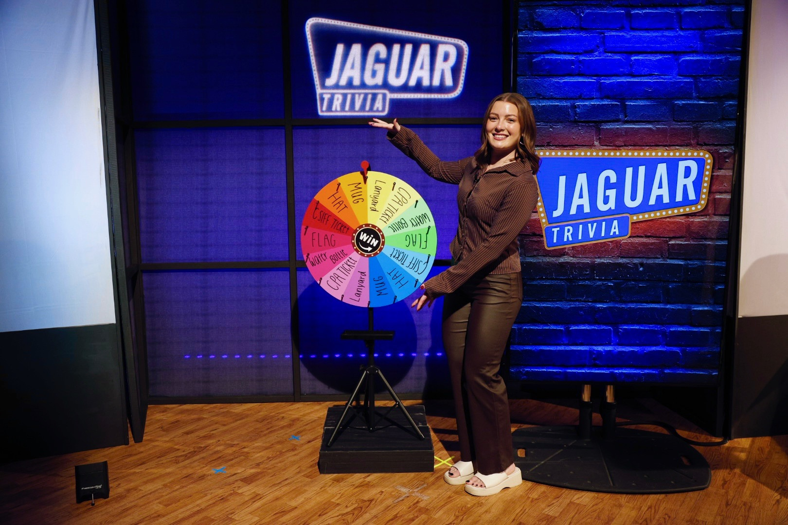 A woman gesturing to a rainbow colored spinning wheel with different trivia subjects on each wedge