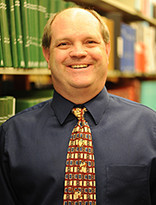 Headshot of a smiling man in shirt and tie standing in front of a shelf of books