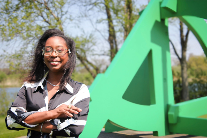 A smiling woman crossing her arms standing in front of a green sculpture on the Governors State University campus