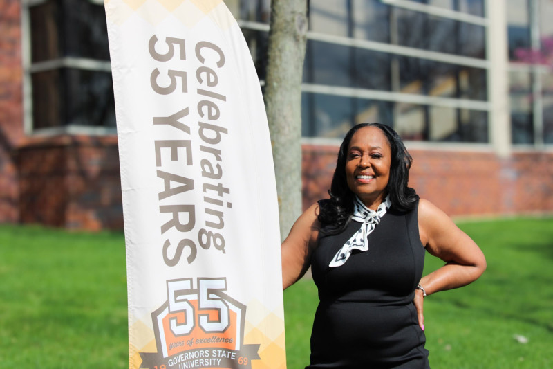 A smiling woman standing outside by a 55 year anniversary flag on the Governors State University campus