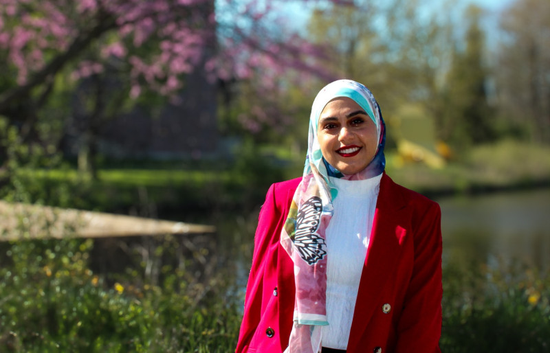 A smiling woman wearing a white shirt, pink blazer, and floral hijab standing outside in front of Governors State University lake