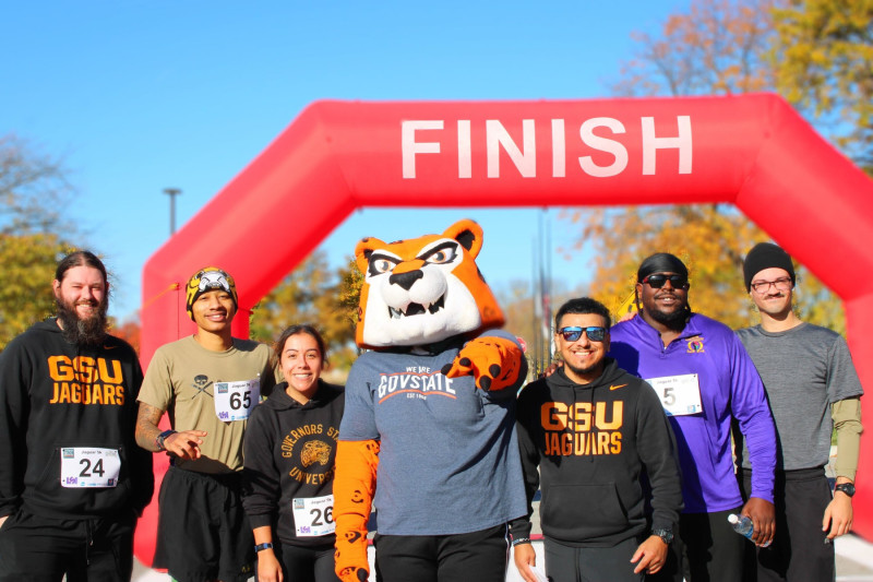 Six people posing with Governors State University mascot Jax the Jaguar at a race finish line