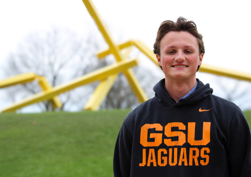 A smiling man wearing a Governors State University hoodie standing in front of a sculpture on campus