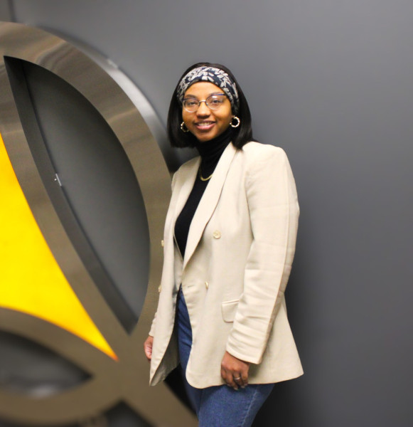 A smiling woman wearing a black shirt and beige blazer standing in front of the Governors State University triad logo in the university library