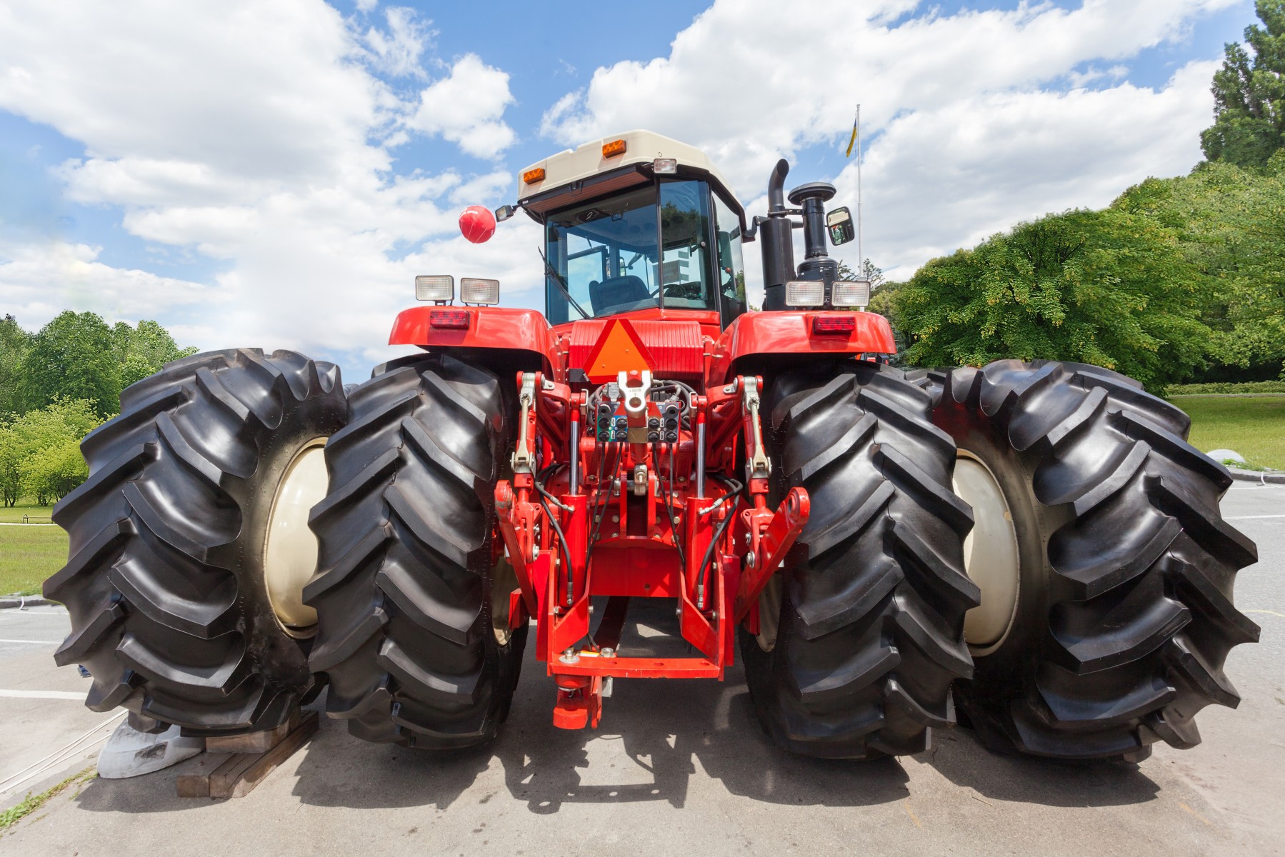 tractor with big wheel and red metal element on back against green tree