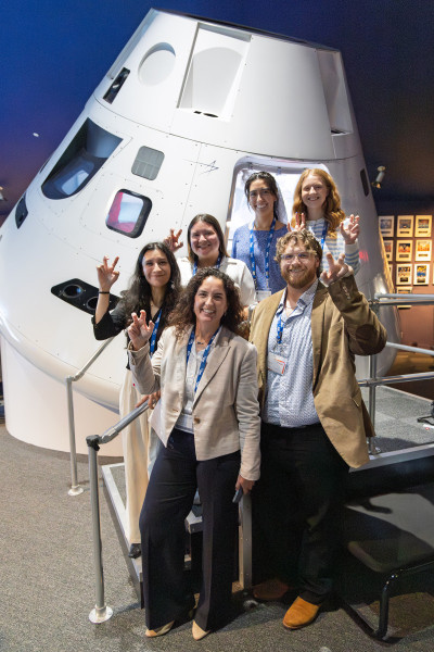 UHCL students pose in front of a spacecraft capsule exhibit during the SHOP-26 summit, smiling and making Hawks hand gestures while wearing conference badges.