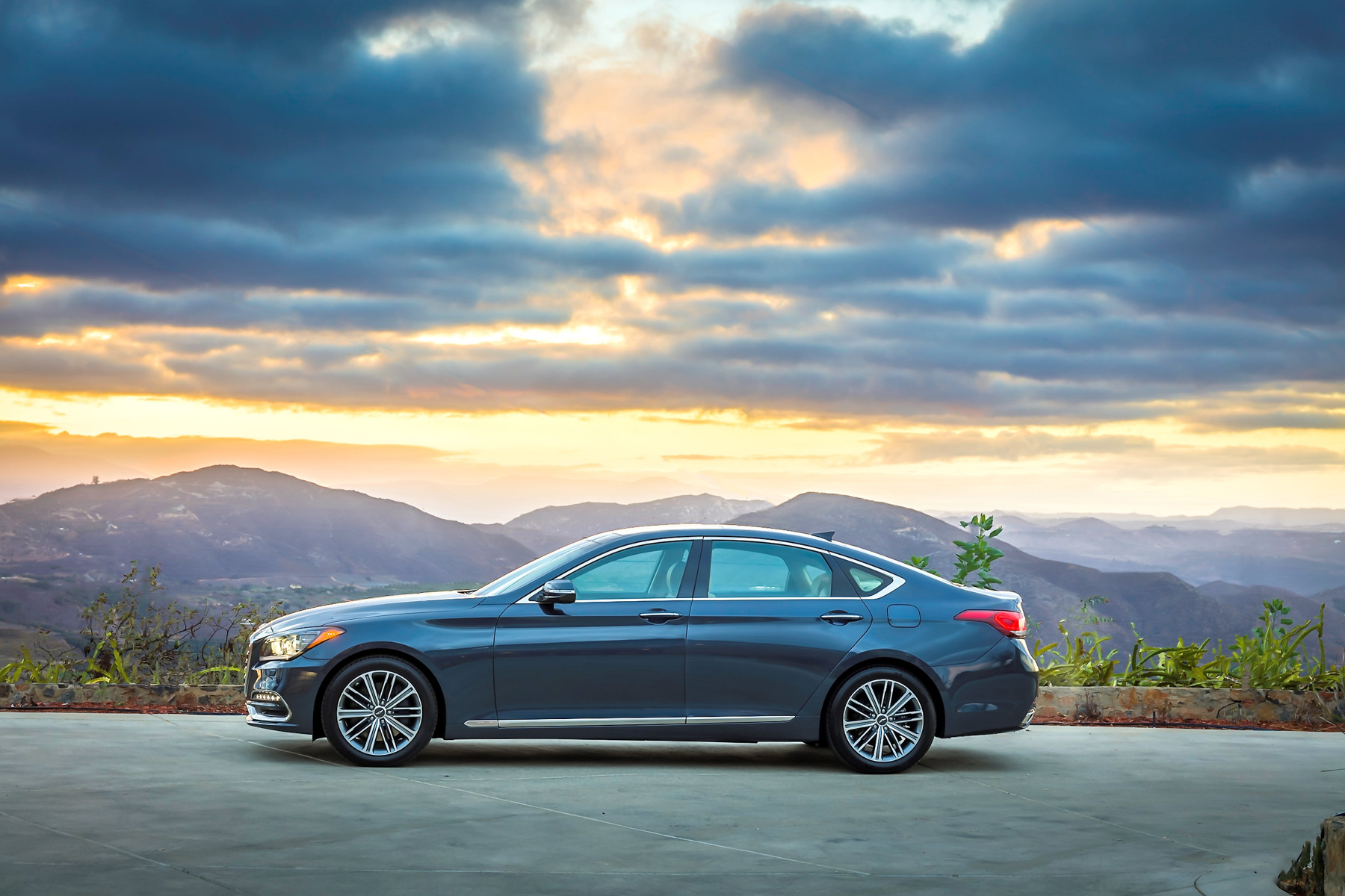 Side view of a dark blue first-generation Genesis G80 sedan parked against a mountain sunset