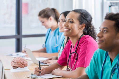 Focus is on a female nursing student as she smiles in the middle of her three classmates as two classmates look forward with her at the professor.  The third classmate looks down and takes notes.  There are windows and a human brain model in the background.