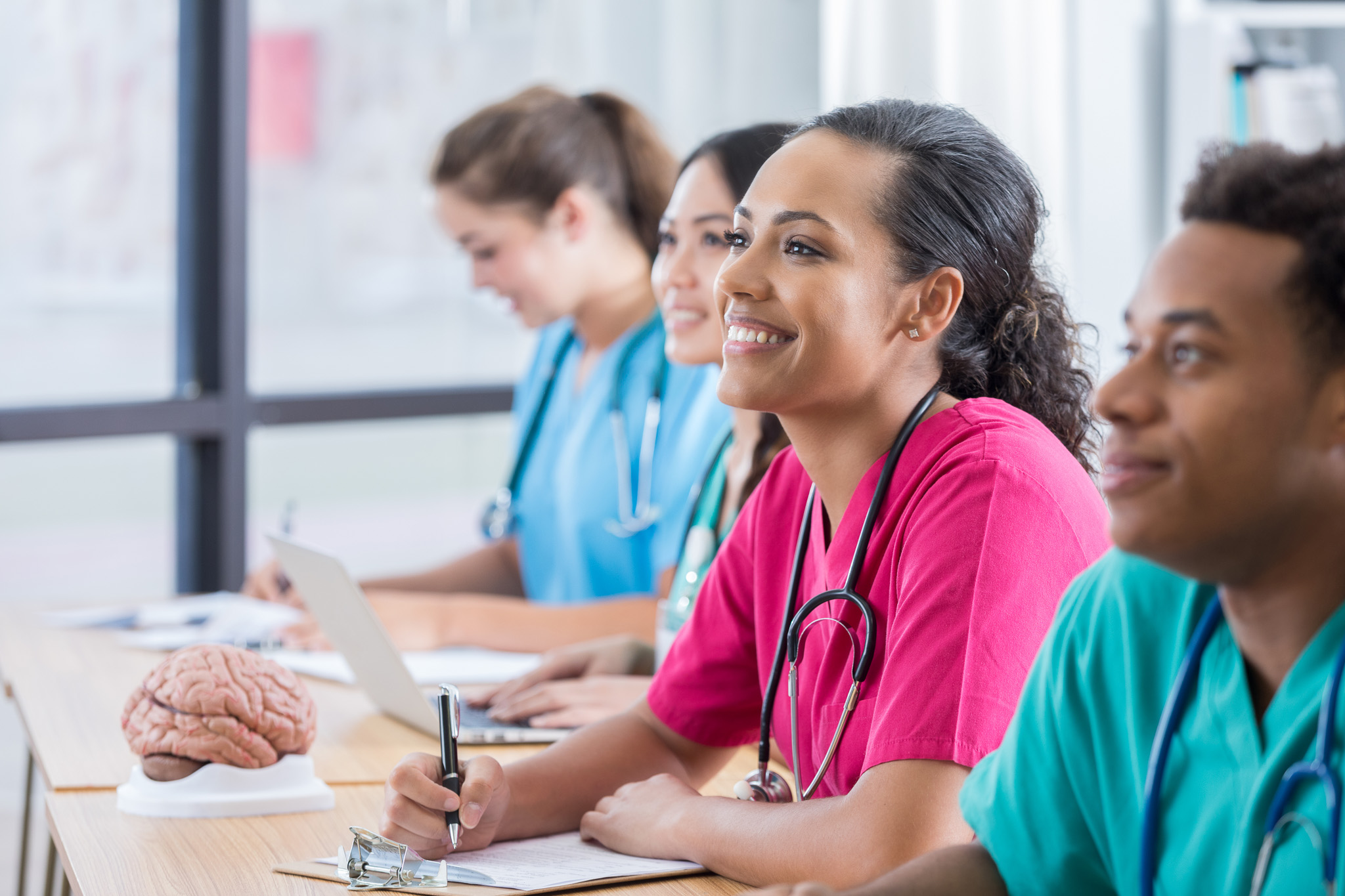 Focus is on a female nursing student as she smiles in the middle of her three classmates as two classmates look forward with her at the professor. The third classmate looks down and takes notes. There are windows and a human brain model in the background.