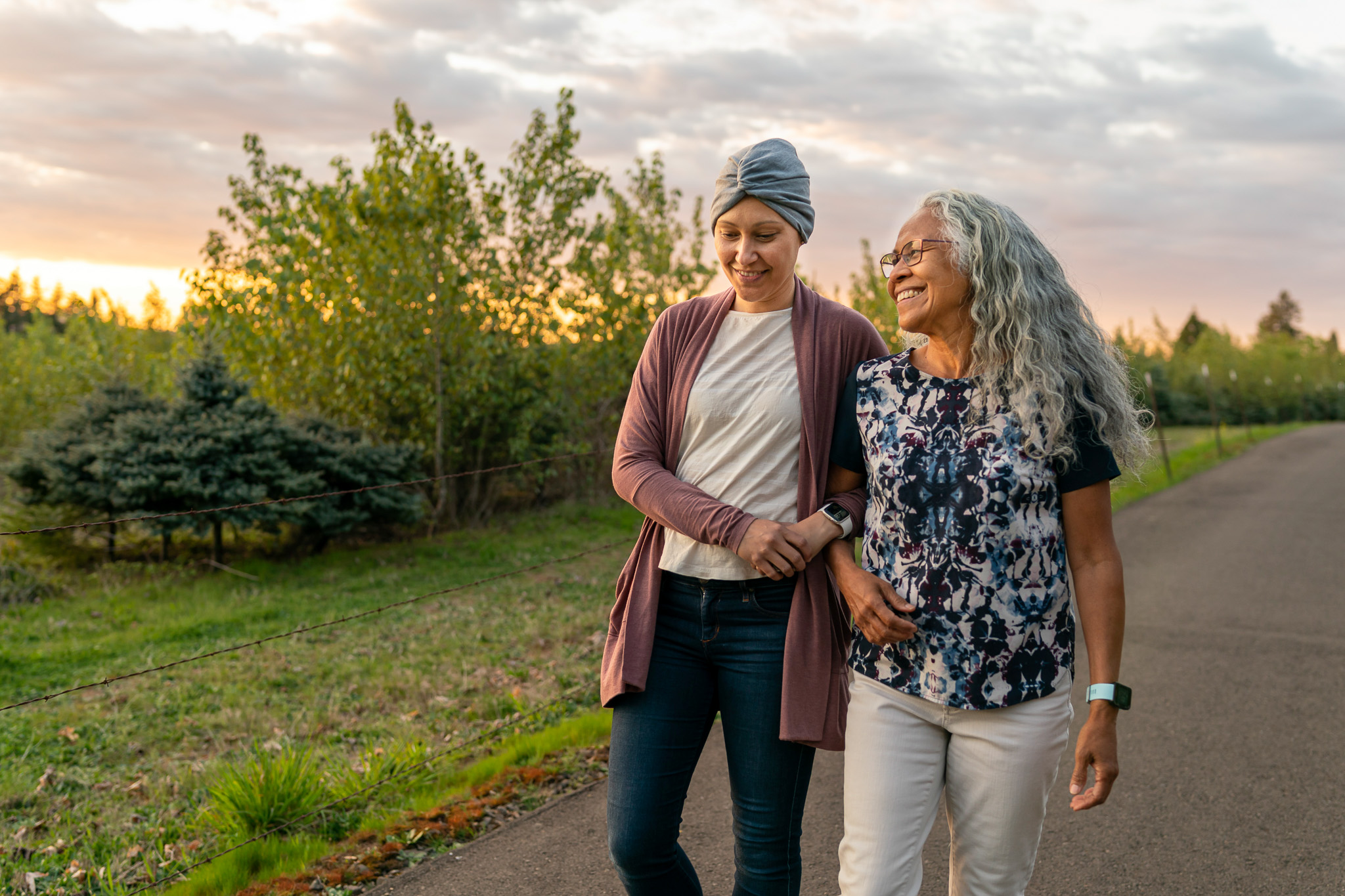 A courageous young mixed race woman recovering from cancer walks arm in arm with her senior mother. They are enjoying the outdoors and time together. They are walking down a rural road at sunset.