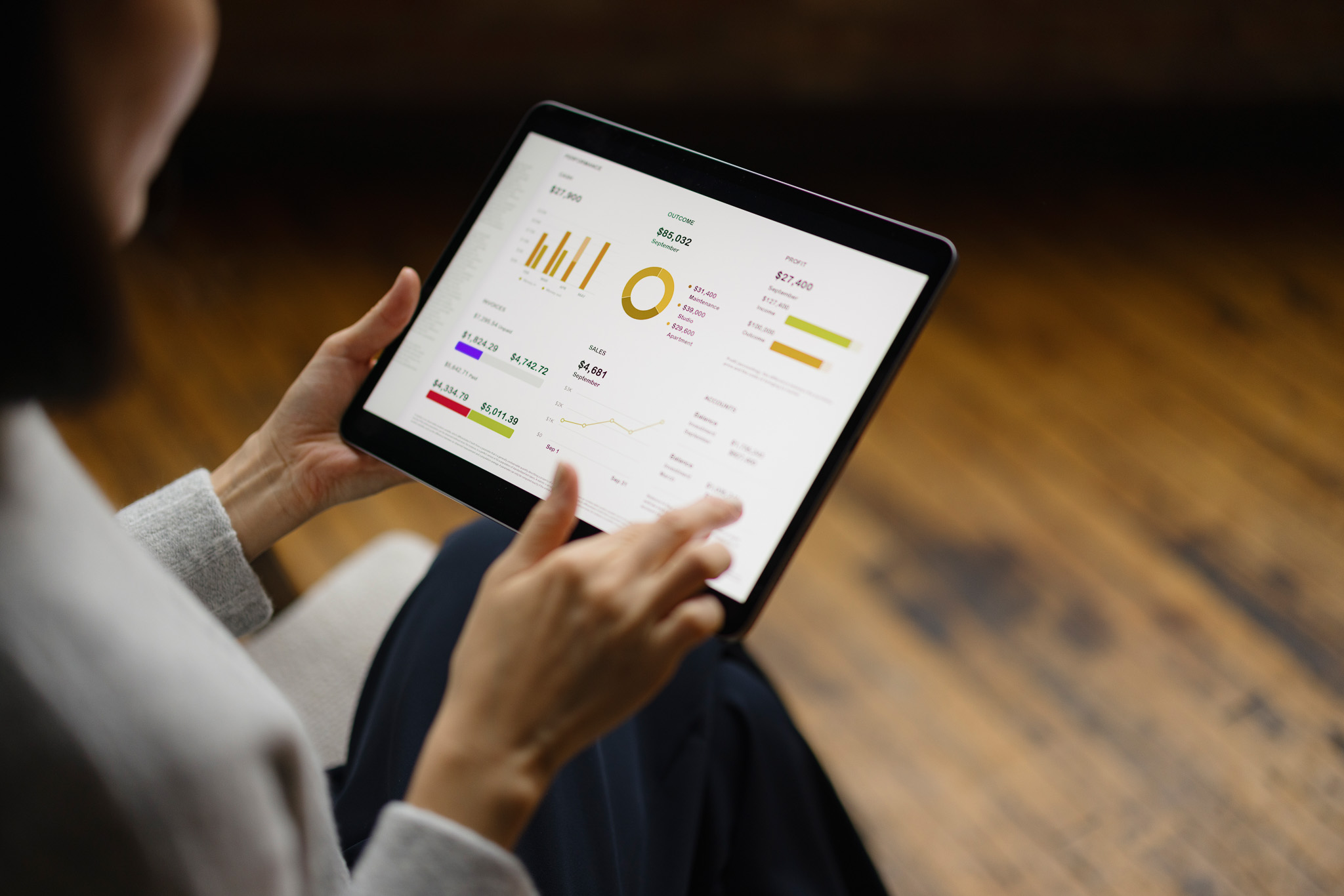 A businesswoman dressed in smart casual attire analyzes digital graphs and charts on a tablet. The setting is a modern office with a warm, wooden floor. Technology aids her business insights.