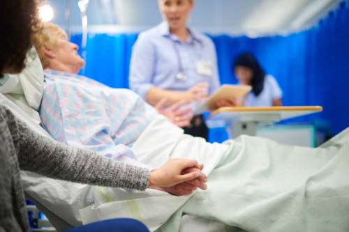 a mid adult female doctor chats to a senior patient on the hospital ward , in the background a young female nurse wearing a hijab is tending to apparatus rack.