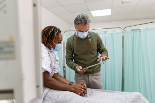 A hospitalized black woman who is being treated for Covid-19 sits in a hospital bed and speaks with her doctor about her recovery. Both individuals are wearing protective face masks. They are reviewing test results on a tablet computer the male doctor is holding.