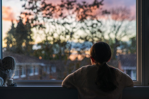 A young girl, with a ponytail, looks out of her bedroom window as the sun is setting. The view through the window is blurry but shows a residential area.