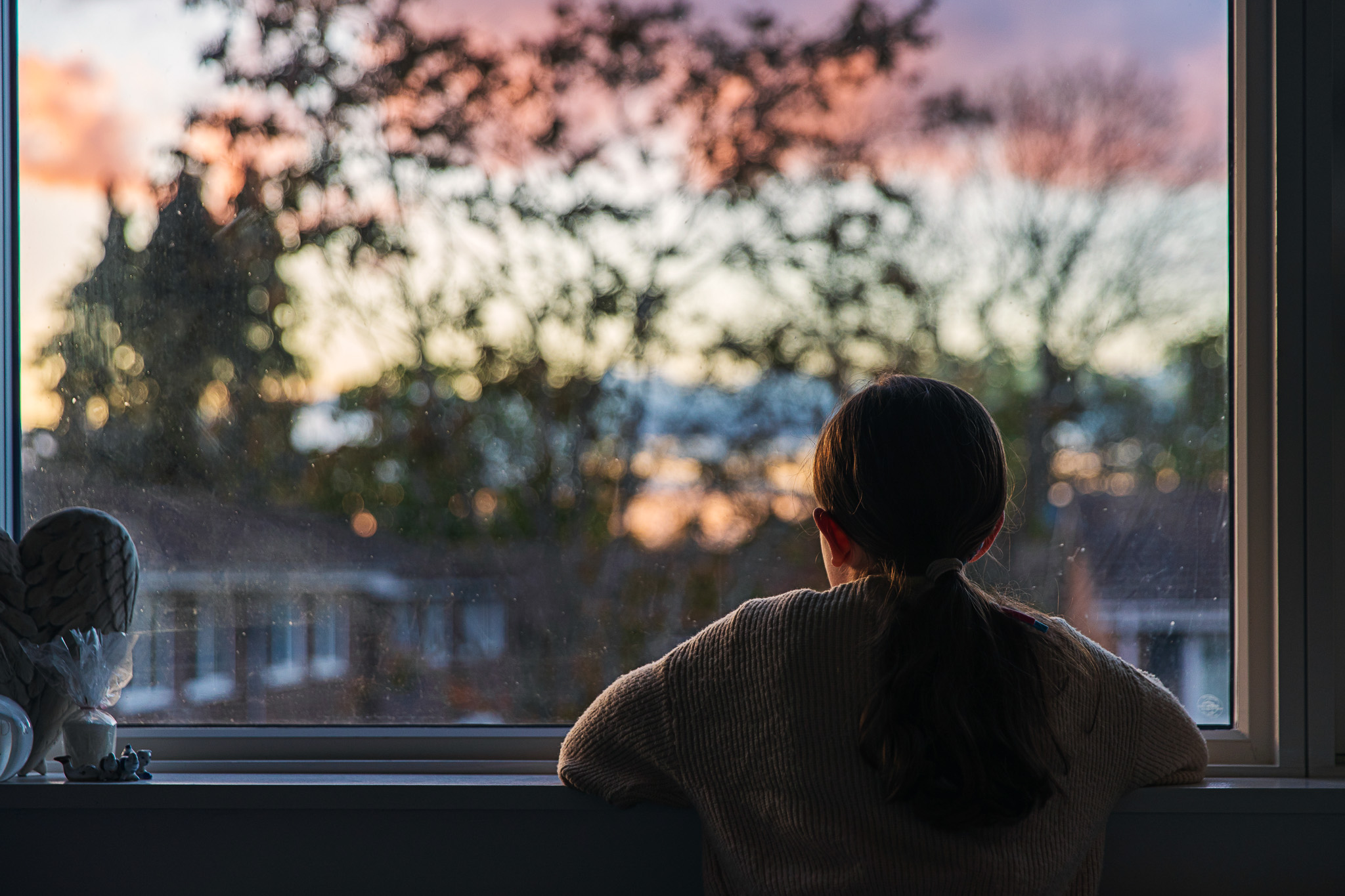 A young girl, with a ponytail, looks out of her bedroom window as the sun is setting. The view through the window is blurry but shows a residential area.