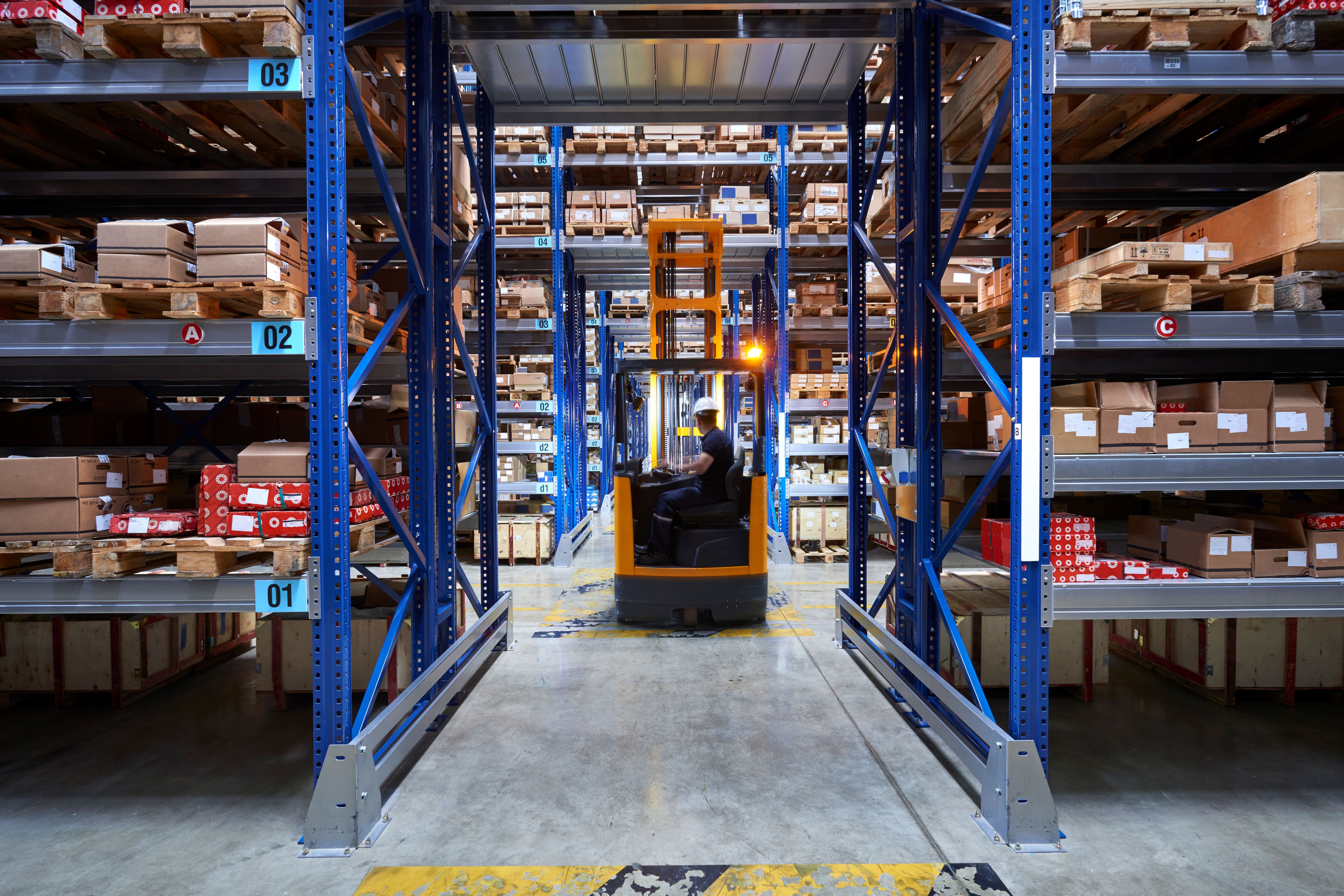Warehouse worker operating a forklift inside a large distribution center with palletized inventory and industrial storage racks supporting freight and logistics operations.