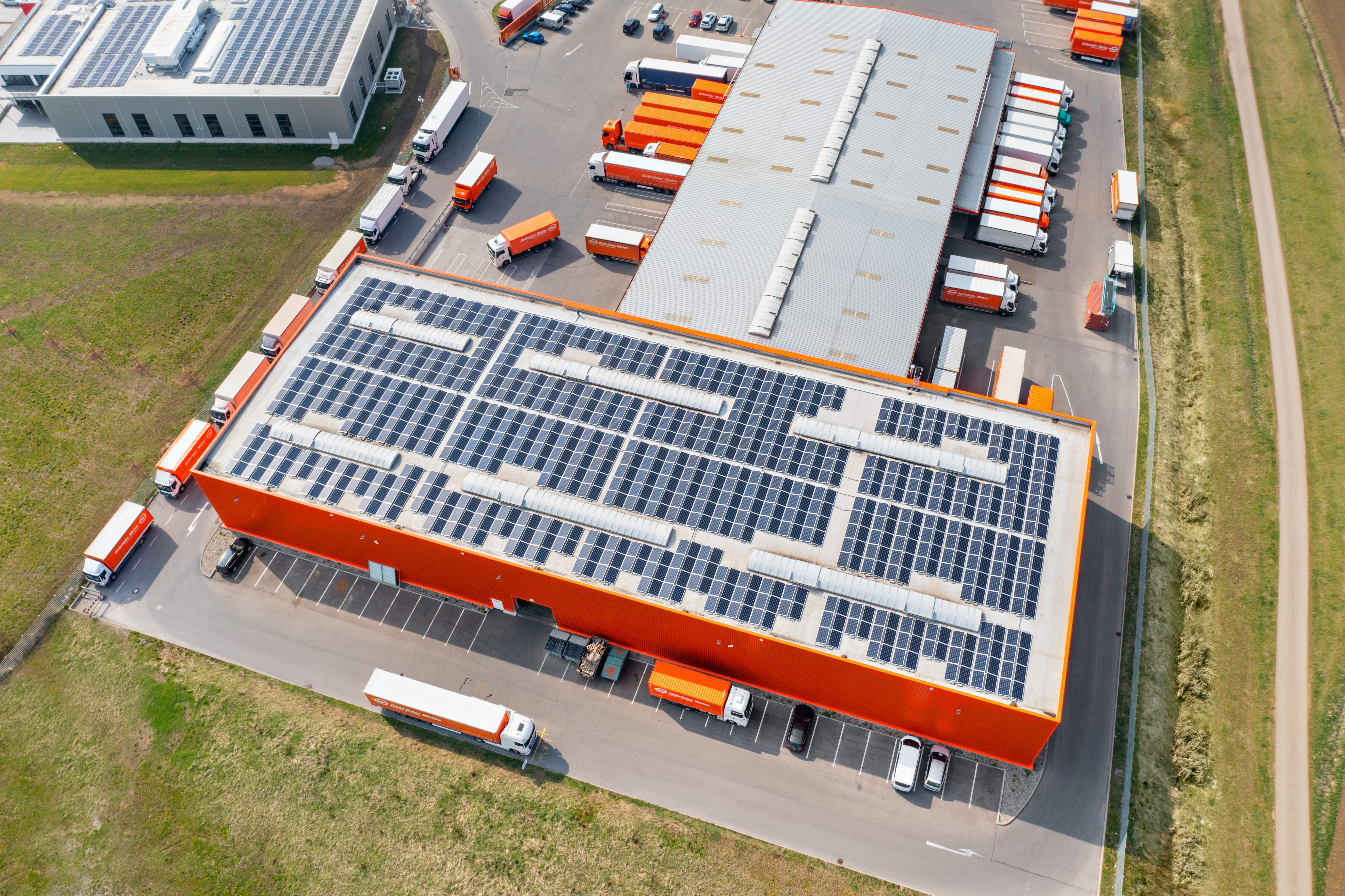 Aerial view of a Gebrüder Weiss logistics facility with rooftop solar panels and orange trucks.