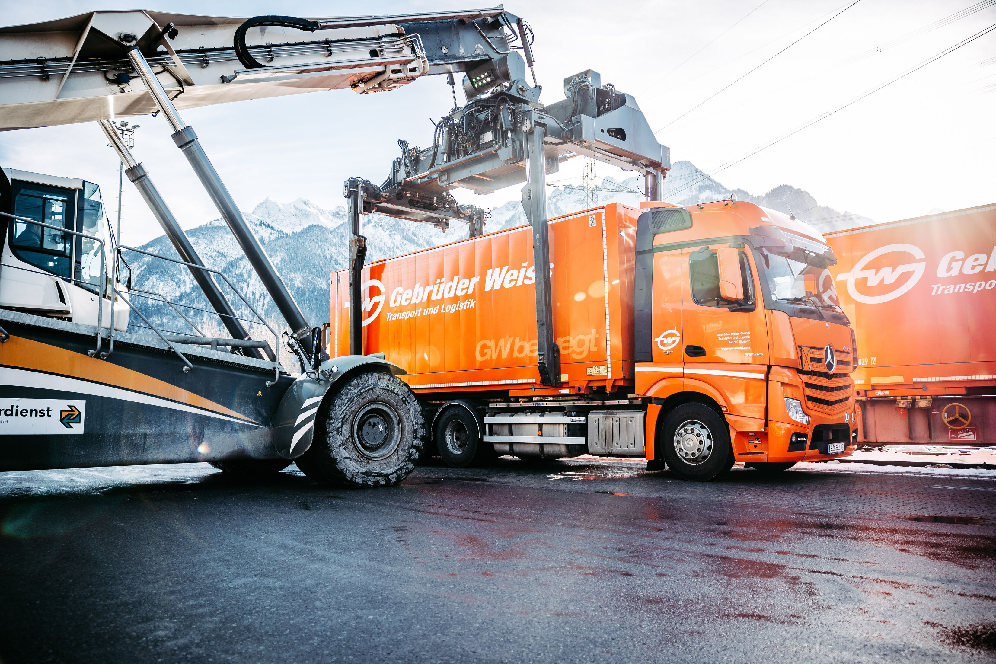 Reach stacker positioned over an orange Gebrüder Weiss container on a truck beside freight railcars.