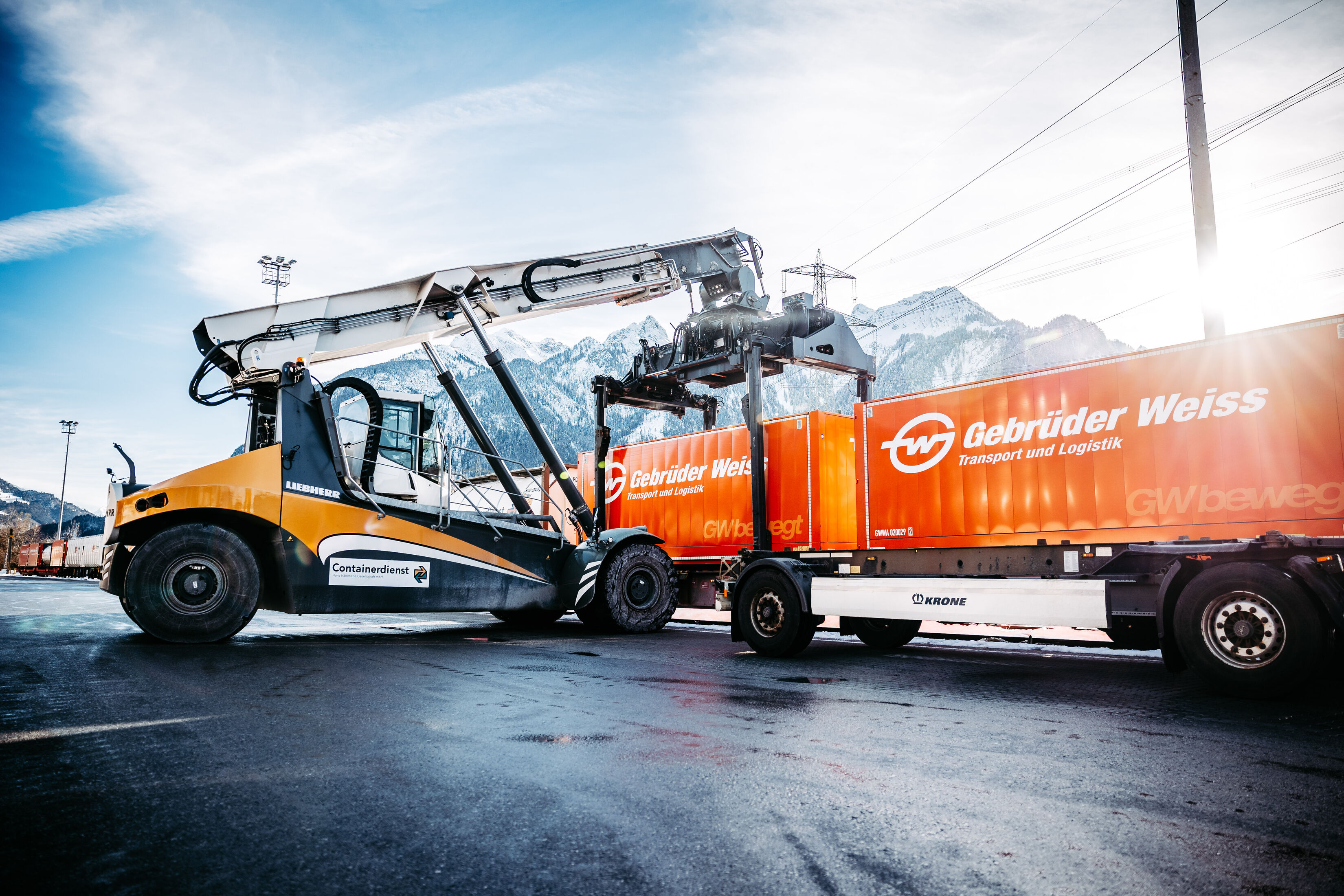 Reach stacker handling an orange Gebrüder Weiss container beside a container trailer at a rail terminal.